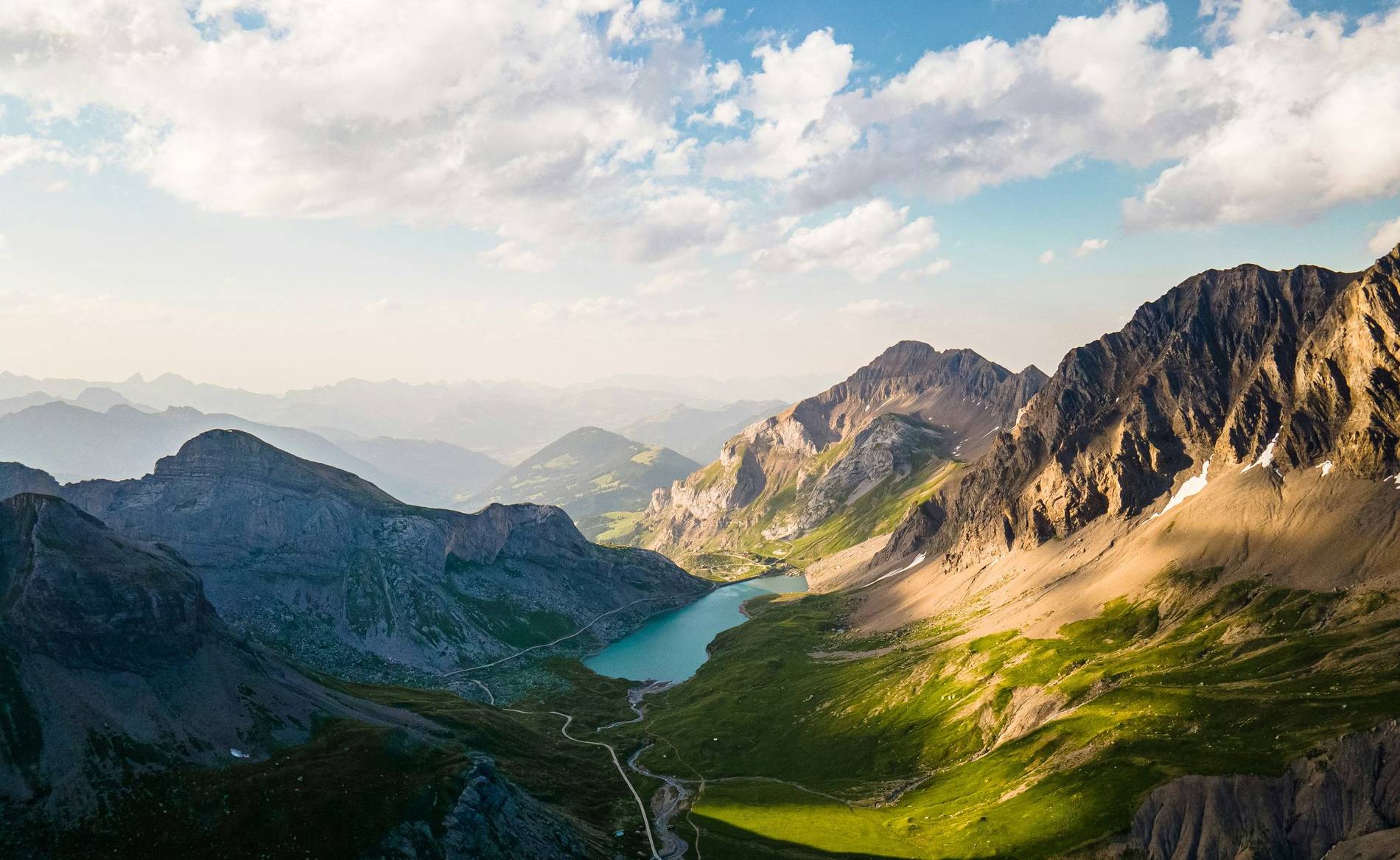 Evening light across an alpine valley with a turquoise reservoir and a pass road climbing away